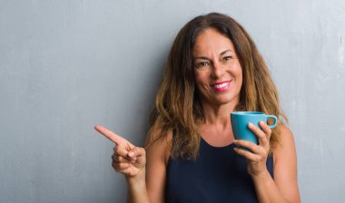 Orta yaş İspanyol kadın kahve el ve parmak ile tarafına işaret çok mutlu içme gri grunge duvarın üzerinden ayakta