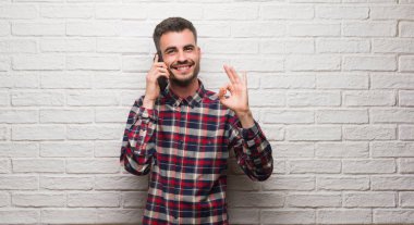 Parmak, mükemmel sembolü ile ok işareti yapıyor beyaz tuğla duvarın üzerinden telefon ayakta konuşan genç yetişkin adam