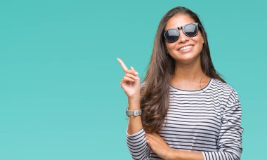 Kameraya bakarak yan el ve parmak ile işaret yüzünde kocaman bir gülümseme ile izole arka plan üzerinde güneş gözlüğü takmış genç güzel Arap kadın.