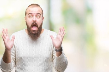 Üzerinde kış kazak giyen genç beyaz hipster adam deli kutluyor arka plan izole ve başarı için kaldırdı kollarını açarak hayrete ve heyecanlı çığlık gözleri açık. Kazanan kavramı