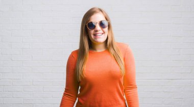 Genç Yetişkin kadın ayakta ve diş gösterilen kendine güvenen bir gülümsemeyle gülümseyerek mutlu bir yüz ile güneş gözlüğü takmış beyaz tuğla duvarın üzerinden