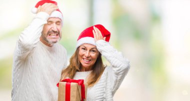 Orta yaş İspanyol çift elle kafasına, utanç ve sürpriz ile şok vurguladı izole arka plan üzerinde Noel şapka ve holding hediye giyen yüz, öfkeli ve sinirli. Korku ve hata için üzgün.