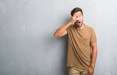 Mahcup ifadeyle parmaklarımın arasından seyir gri grunge duvar yüz ve gözler el ile kapsayan şok bakmak üzerinde yakışıklı genç adam.