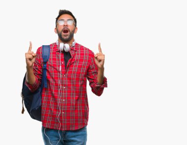 Kulaklıklar ve sırt çantası izole arka plan üzerinde giyen yetişkin İspanyol öğrenci adam hayret ve yukarıya bakıp parmakları ile işaret şaşırttı ve silah kaldırdı.