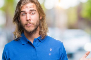 Yakışıklı delikanlı izole arka plan clueless ve şaşkın bir ifadeyle kollar ve eller kaldırdı üzerinde uzun saçlı. Şüphe kavramı.