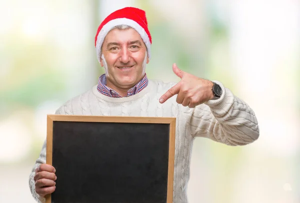 英俊的老人戴着圣诞帽子和持有黑板在孤立的背景非常高兴地指着手和手指 — 图库照片