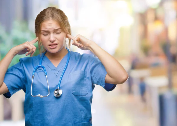 Young caucasian doctor woman wearing surgeon uniform over isolated background covering ears with fingers with annoyed expression for the noise of loud music. Deaf concept.