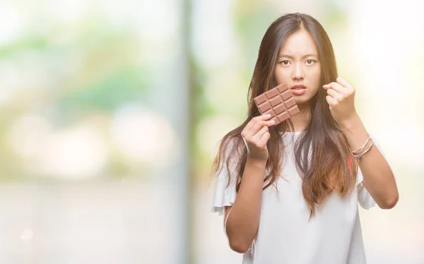 Young asian woman eating chocolate bar over isolated background annoyed and frustrated shouting with anger, crazy and yelling with raised hand, anger concept