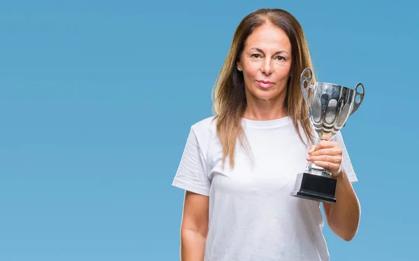 Middle age hispanic winner woman celebrating award holding trophy over isolated background with a confident expression on smart face thinking serious