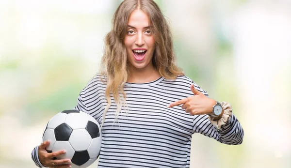 美丽的年轻金发女子拿着足球在孤立的背景与惊喜的脸指向自己的手指 — 图库照片