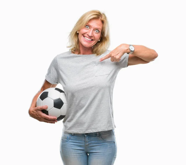 中年金发女子持有足球足球球超过孤立背景与惊讶的脸指向自己的手指 — 图库照片