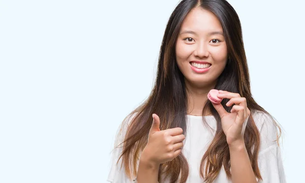笑顔の サイン 優秀な兆候を親指をやって幸せな孤立の背景の上甘いピンクのマカロンを食べる若いアジア女性 — ストック写真