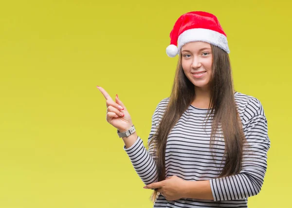 若い美しい白人女性の顔に大きな笑みを浮かべて隔離された背景にクリスマスの帽子を着てカメラを見て側の手と指で指す — ストック写真