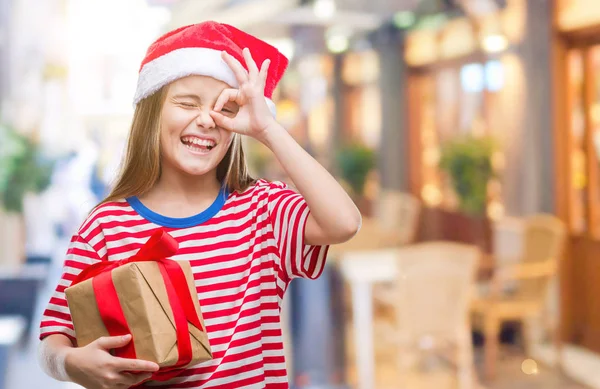 指を通して見る目に手で サインをして笑っている幸せそうな顔で孤立した背景にクリスマスの帽子 持株ギフトを着た美しい少女 — ストック写真