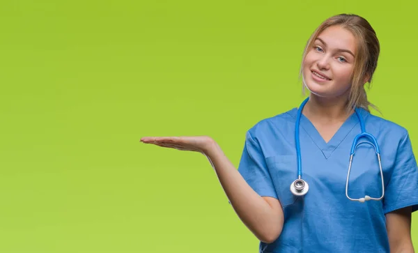 年轻的白种人医生妇女穿外科医生制服在孤立的背景微笑欢快的呈现和指向手掌的手看着相机 — 图库照片