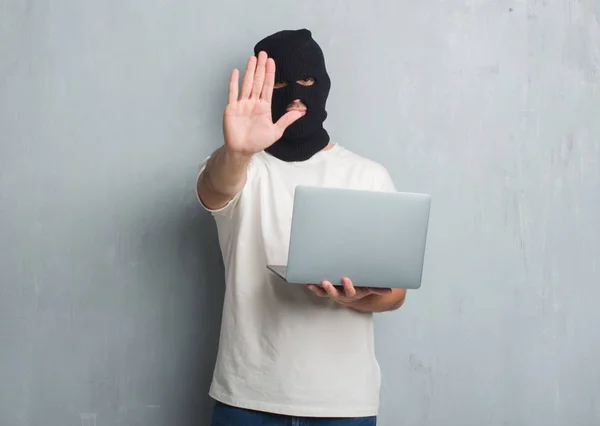 Young caucasian hacker man over grey grunge wall doing cyber attack using laptop with open hand doing stop sign with serious and confident expression, defense gesture