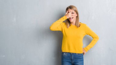 Mahcup ifadeyle parmaklarımın arasından seyir grunge gri duvar yüz ve gözler el ile kapsayan şok bakmak üzerinde duran güzel genç kadın.