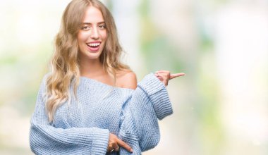 Kameraya bakarak yan el ve parmak ile işaret yüzünde kocaman bir gülümseme ile izole arka plan üzerinde kış kazak giyen güzel sarışın genç kadın.