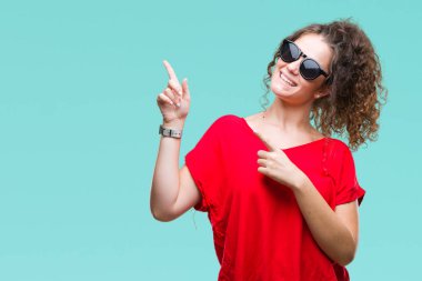Güzel esmer kıvırcık saçlı Genç kız gülümseyerek ve iki elleri ve parmakları ile tarafına işaret kamera bakarak izole arka plan üzerinde güneş gözlüğü takıyor.