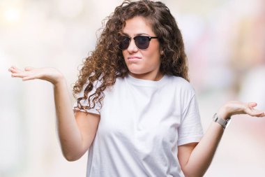 Güzel esmer kıvırcık saçlı Genç kız üzerinde güneş gözlüğü takıyor arka plan clueless ve şaşkın bir ifadeyle kollar ve eller kaldırdı izole. Şüphe kavramı.