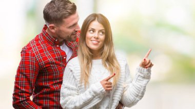 Gülümseyen ve iki elleri ve parmakları ile tarafına işaret kamera bakarak izole arka plan üzerinde kış kazak giyen aşık genç çift.