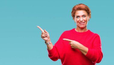 Gülümseyen ve iki elleri ve parmakları ile tarafına işaret kamera bakarak izole arka plan üzerinde kış kazak giyen Atrractive üst düzey beyaz Kızıl saçlı kadın.