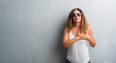 Orta yaş İspanyol kadın gri duvar kapalı gözler ile göğüs ellerinde ve yüzünde minnettar jest ile gülümseyen güneş gözlüğü takıyor. Sağlık kavramı.