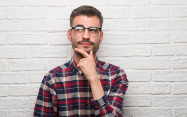 Genç yetişkin adam beyaz tuğla duvar ciddi bir yüz düşünme hakkında soru, çok karışık bir fikir üzerinde ayakta