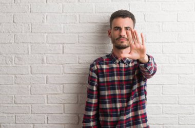 Genç yetişkin adam dur yapıyor beyaz tuğla duvardan ayakta elin avuç ile şarkı. Negatif ve ciddi bir jest yüz ifadesiyle uyarı.