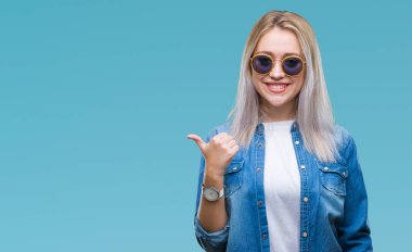 Genç Sarışın kadın arıyorum ve başparmak yanında yukarıyı mutlu yüz ile gülümseyen izole arka plan üzerinde güneş gözlüğü takıyor.