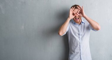 Genç redhead iş adamı, dilini parmaklarının arasından baktığını yapışmasını dürbün gibi ok jest yapıyor gri grunge duvarın üzerinden. Deli ifade.