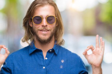 İzole arka plan üzerinde güneş gözlüğü takmış uzun saçlı yakışıklı delikanlı rahatla ve gülümseyen gözlerle kapalı parmak jestle meditasyon yapıyor. Yoga kavramı.