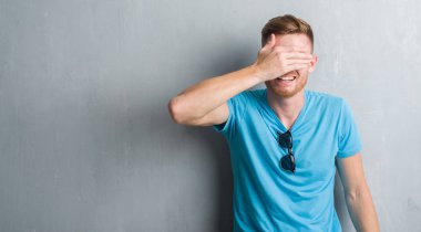 Gri grunge duvar rahat kıyafet gülümseyen ve gözler için sürpriz kapsayan yüz el ile gülüyor giyen genç Kızıl saçlı adam. Kör kavramı.