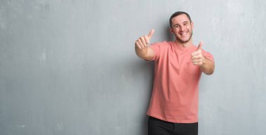 Gri grunge duvar onaylama üzerinde beyaz delikanlı el ile pozitif jest yapıyor gülümseyen ve başarı için mutlu yaşasın. Kamera, kazanan jest arıyorsunuz.