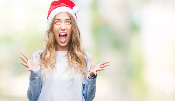 美丽的年轻金发女子戴着圣诞节帽子在孤立的背景疯狂和疯狂的呼喊和咆哮与积极的表达和武器提出 挫折概念 — 图库照片