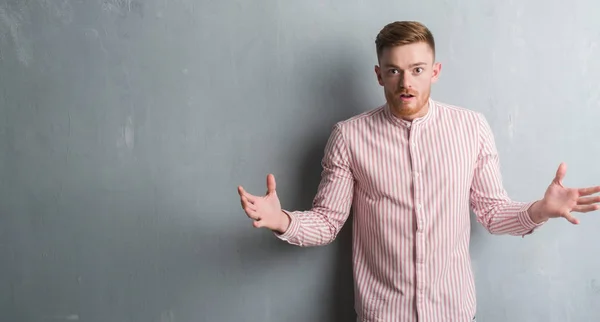Young redhead man over grey grunge wall scared in shock with a surprise face, afraid and excited with fear expression
