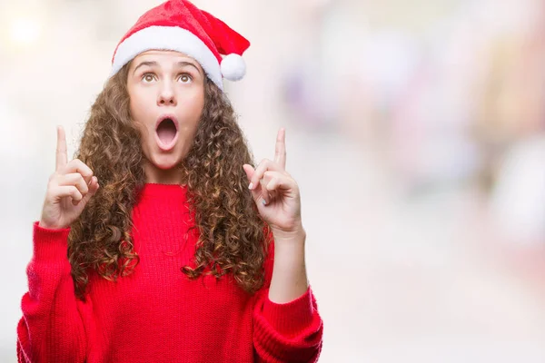 孤立した背景にクリスマスの帽子をかぶって若いブルネットの少女びっくり驚いて見上げると指で指していると腕を上げた — ストック写真