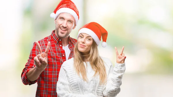 勝利のサインを行うカメラでウィンクしている幸せそうな顔を浮かべて隔離された背景にクリスマスの帽子をかぶっての愛の若いカップル — ストック写真