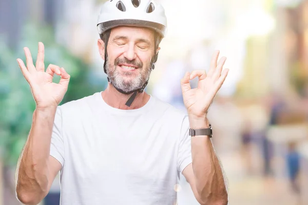 Middle age senior hoary cyclist man wearing bike safety helment isolated background relax and smiling with eyes closed doing meditation gesture with fingers. Yoga concept.