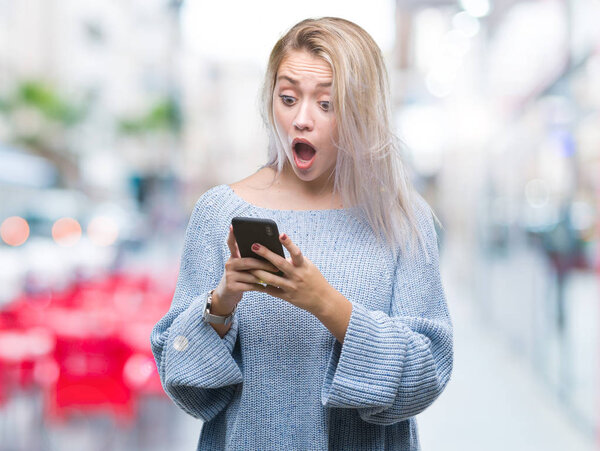Молодая блондинка пишет смс с помощью смартфона на изолированном фоне, напуганная от удивления лицом, напуганная и взволнованная выражением страха
