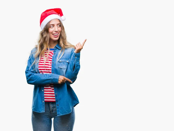 美しい若いブロンドの女性の顔に大きな笑みを浮かべて隔離された背景にクリスマスの帽子を着てカメラを見て側の手と指で指す — ストック写真