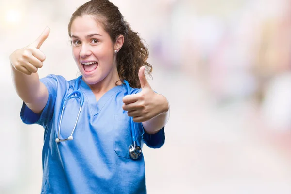 Young brunette doctor girl wearing nurse or surgeon uniform over isolated background approving doing positive gesture with hand, thumbs up smiling and happy for success. Looking at the camera, winner gesture.