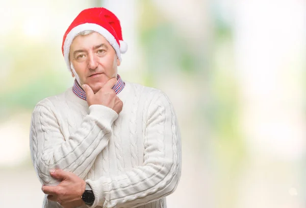 ハンサムな年配の男性が組んだ腕と手をあごに上げた笑顔でカメラに自信を持って探して孤立の背景にクリスマス帽子をかぶっています 前向きに考えてください — ストック写真