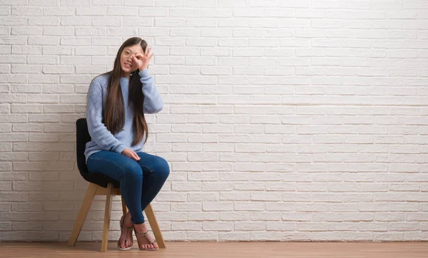 Young Chinese woman sitting on chair over brick wall with happy face smiling doing ok sign with hand on eye looking through fingers