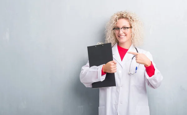 Jovem Mulher Médica Loira Sobre Grunge Parede Cinza Segurando Prancheta — Fotografia de Stock