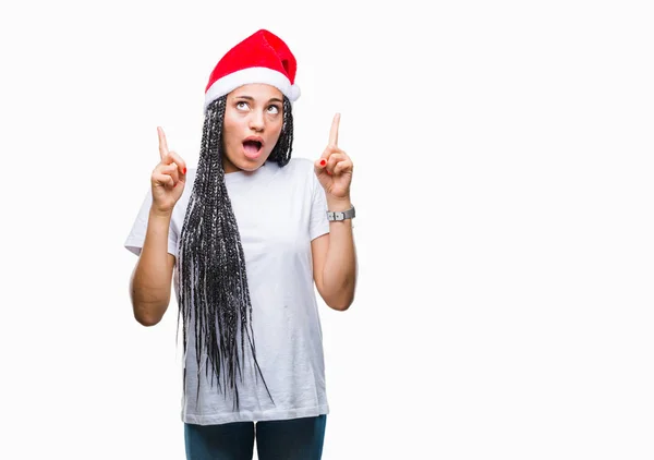 年轻的编织头发非洲裔美国女孩戴着孤立的背景圣诞帽惊讶和惊讶地抬起头来 用手指指指点点 举起双臂 — 图库照片