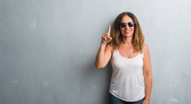 Orta yaş İspanyol kadın gri duvar gösterilen ve parmak numara ile yukarı dönük kendinden emin ve mutlu gülümseyen güneş gözlüğü takıyor.