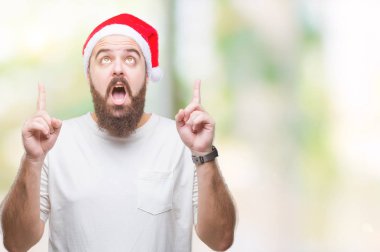 Giyen Noel şapka izole arka plan üzerinden hayran ve yukarıya bakıp parmakları ile işaret şaşırttı ve silah kaldırdı Genç beyaz hipster adam.