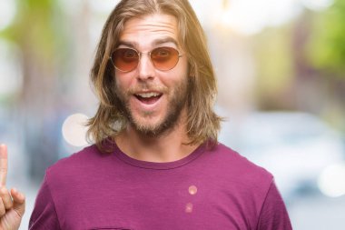 Genç yakışıklı adam uzun saçlı başarılı fikirle izole arka plan işaret parmağı üzerinde güneş gözlüğü takıyor. Çıktıktan ve mutlu. Bir numara.