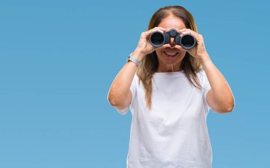 Orta yaş İspanyol kadın ayakta ve diş gösterilen kendine güvenen bir gülümsemeyle gülümseyerek mutlu bir yüz ile izole arka plan üzerinde dürbün arıyor